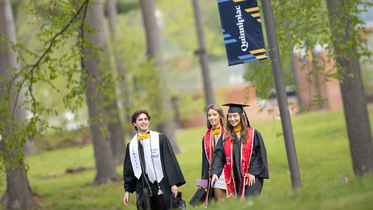 Three graduates walk under a Quinnipiac banner