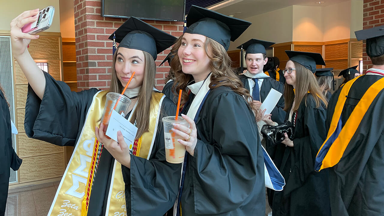 Graduates pose for a selfie while holding their iced coffees while lining up for procession