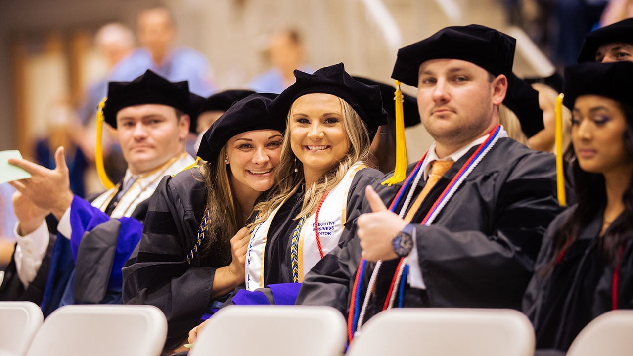 Graduates sit in their chairs and smile with thumbs up