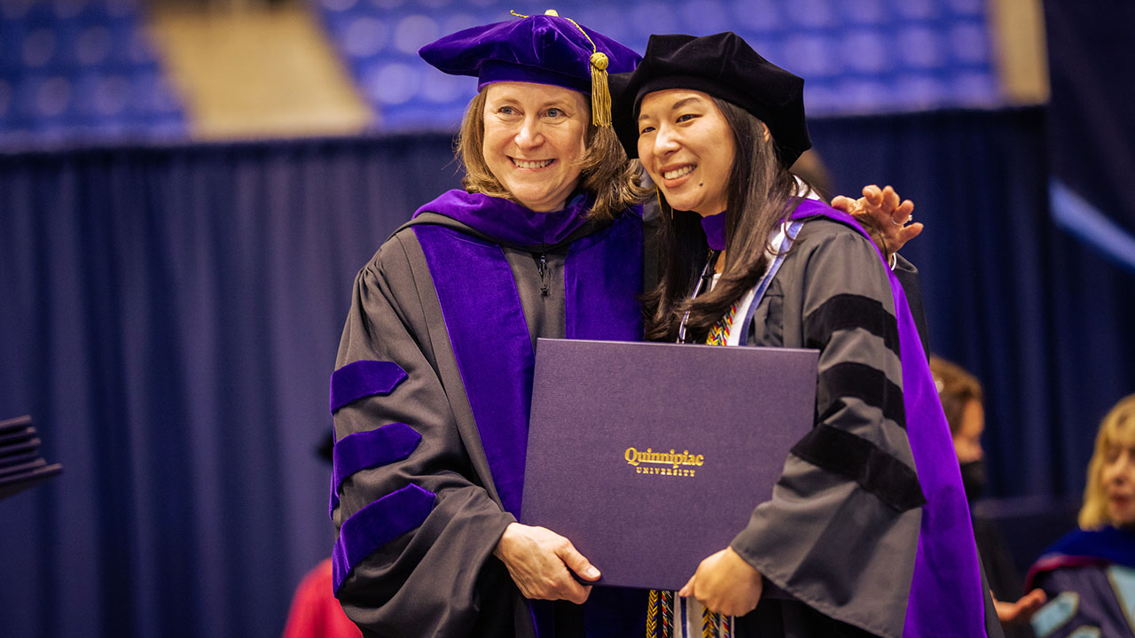 A graduate and professor pose with diploma and smile at a camera
