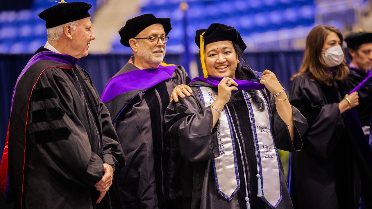 A graduate fixes her hood on stage with help from professors behind her