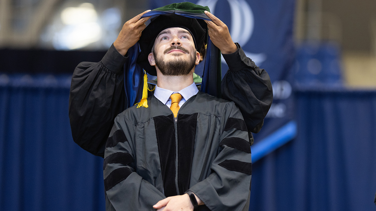 A graduate stands looking up in admiration as he is hooded on stage