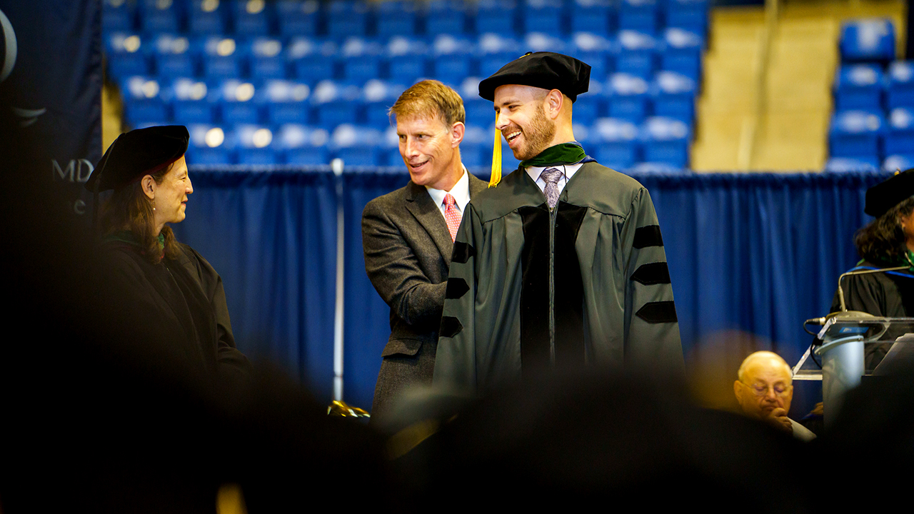 A graduate smiles at their professor on stage after being hooded by a relative behind them
