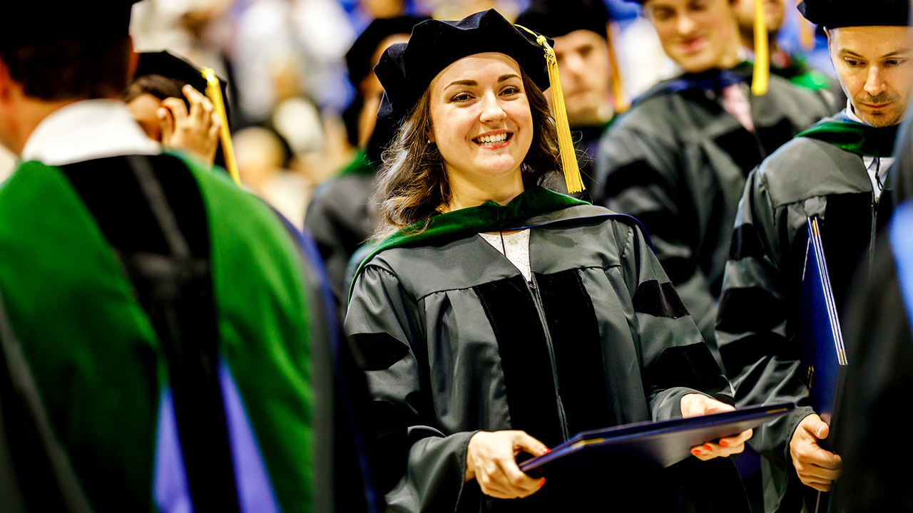 A graduate smiles holding her diploma after being hooded.