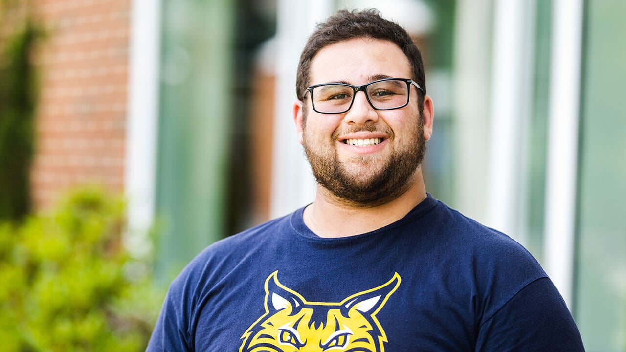 paul capuzzo poses smiling in a blue shirt with the quinnipiac bobcat on it