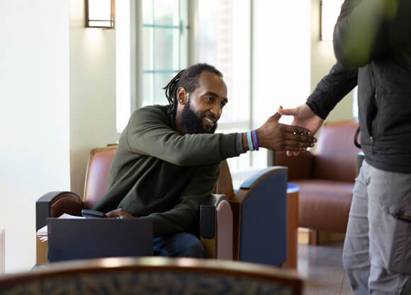 A male student shakes the hand of another student who is out of frame while sitting with his laptop