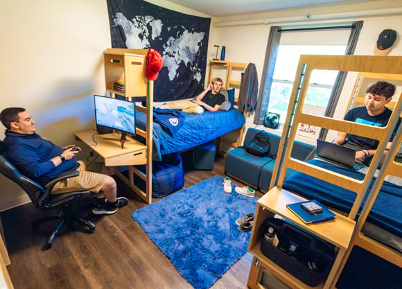 Two Quinnipiac students lounge on their beds with computers and one plays a computer game