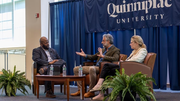 Three people sit in upholstered chairs and talk together in front of a Quinnipiac banner