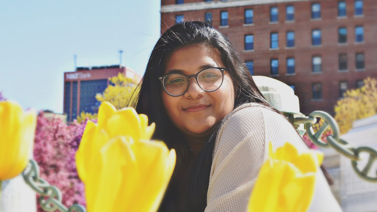 A student smiling at the camera, crouching behind the foreground of yellow flowers. They are wearing glasses and have long, dark hair.