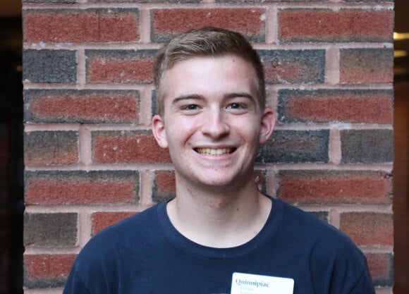 Student leaning against a brick wall, smiling at the camera. They are wearing a Resident Assistant name tag on their shirt.