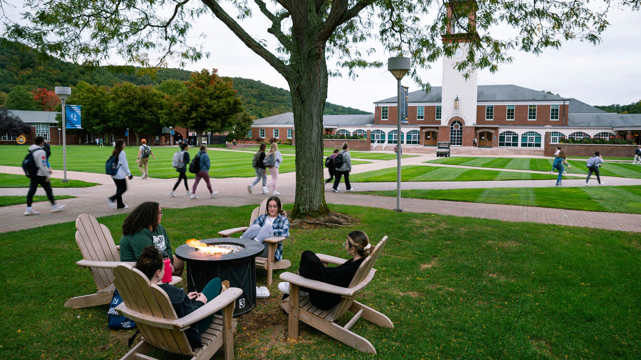 Students sitting in wooden chairs by the fire on the quad as students walk by