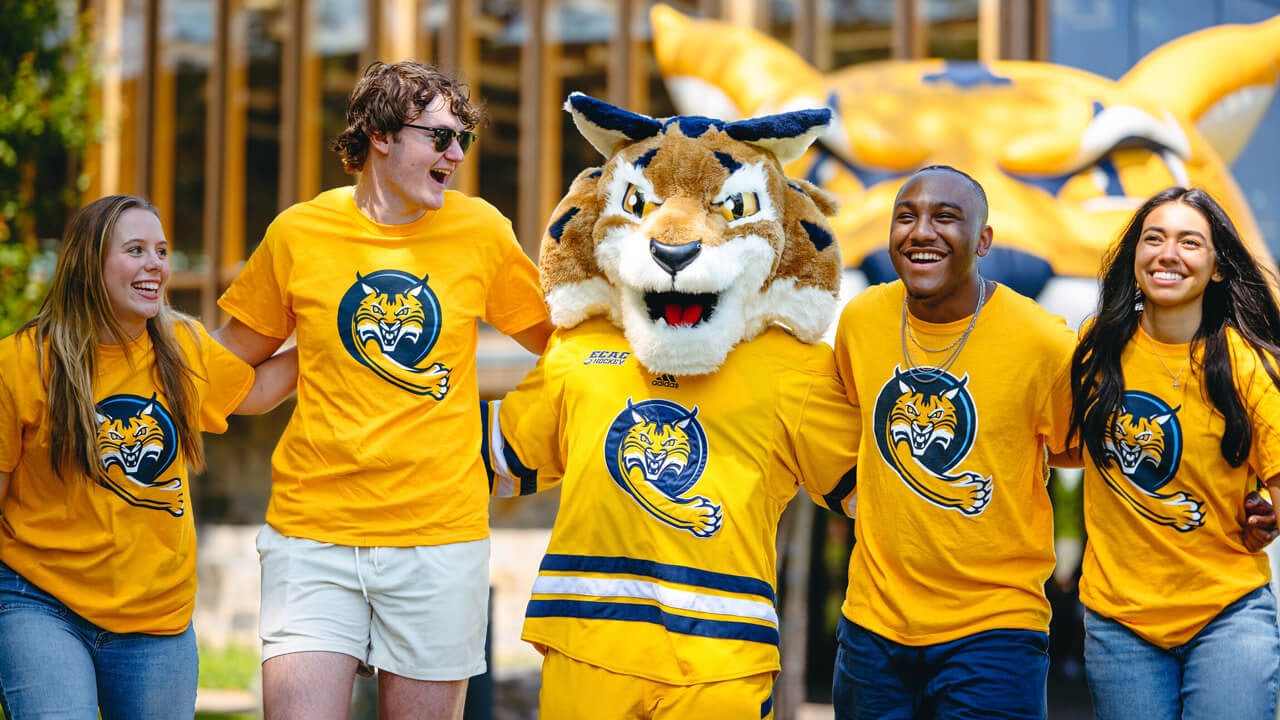 Four students and the bobcat mascot lock arms and walk together wearing matching shirts