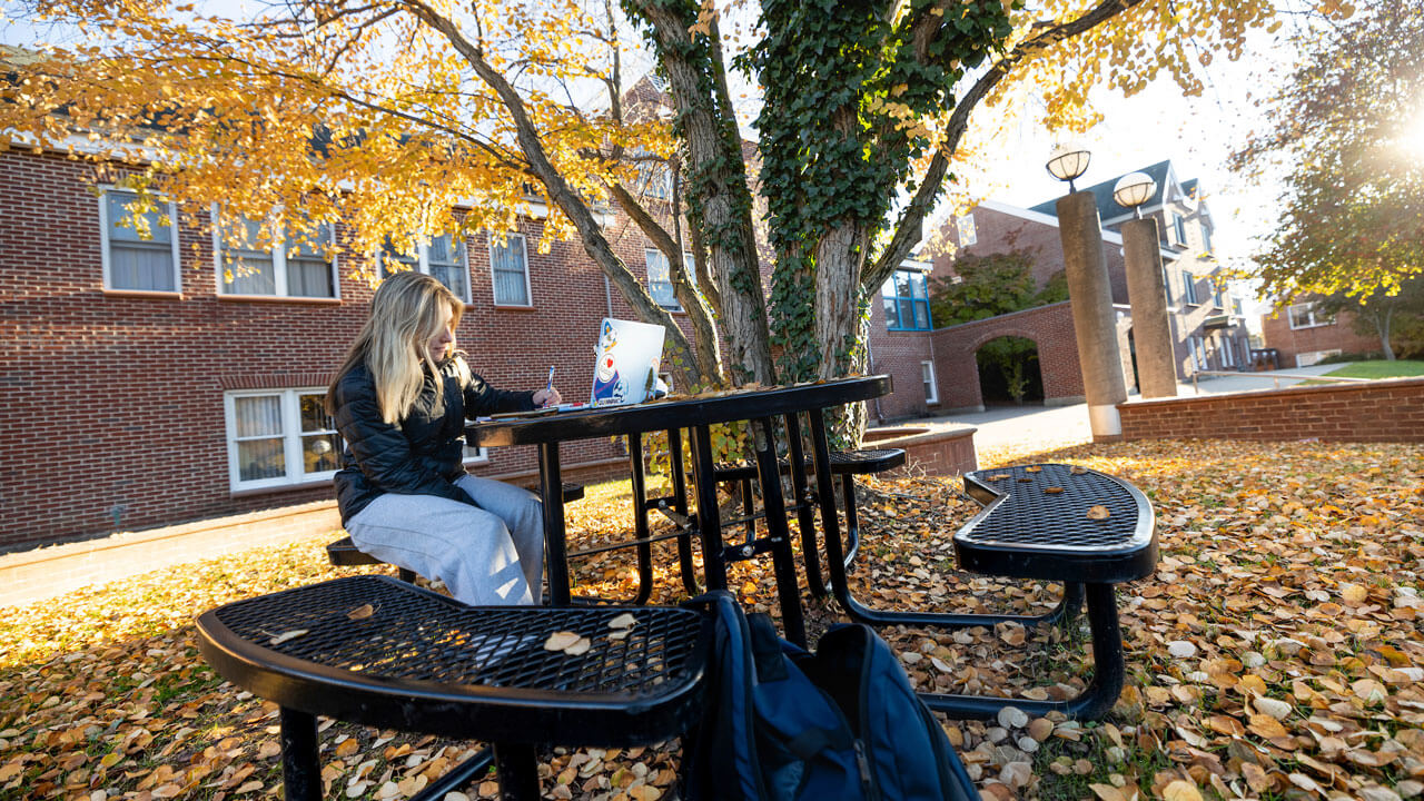 Student sitting at table by Village with the fall foliage surrounding her