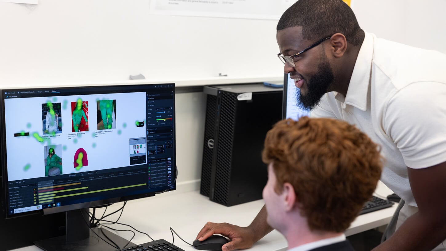 A student and faculty member examine heat map data on an eye-tracking interface displayed on a desktop monitor. The screen shows visual analytics for user engagement with a product webpage, while the faculty member smiles and provides guidance.
