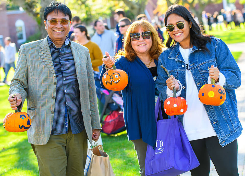 A Quinnipiac student smiles and poses with her parents holding painted pumpkins during a family event