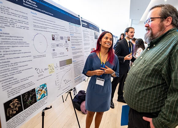 Nicole Wijendra looking at her poster and speaker while a man looks at the poster