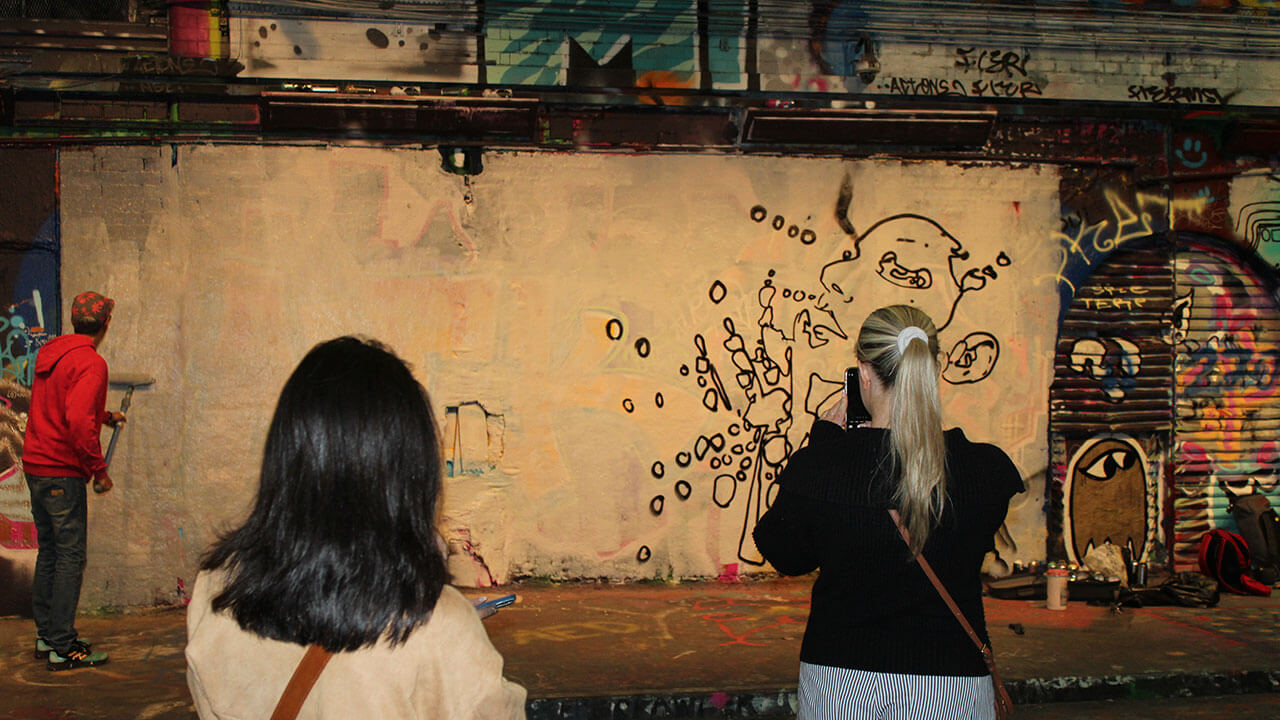 two students take pictures of a man adding graffiti to a tunnel wall