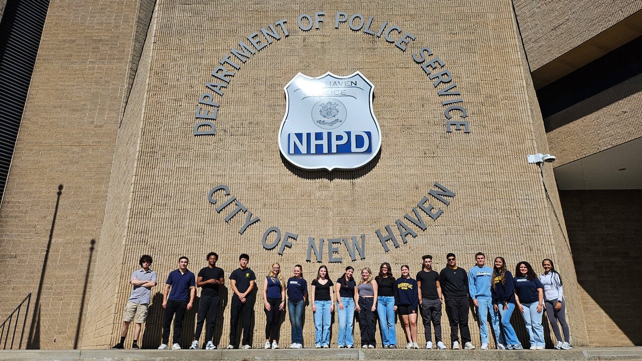 A group of 17 students stands outside the New Haven Police Department building, underneath the logo on the wall, at the top of a staircase.