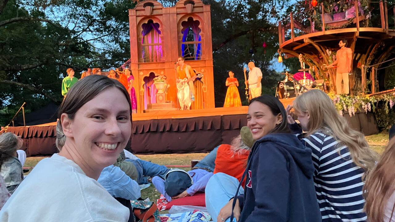 A seated student and faculty member looking back at the camera while performers in colorful costumes play musical instruments onstage.