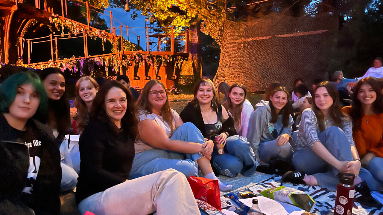 A group of 10 students sitting on a picnic blanket with a backdrop of an outdoor stage set.