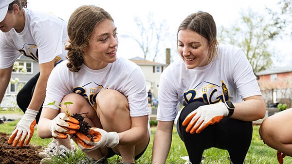 two female students wear gardening gloves and smile, one holds a small plant in dirt