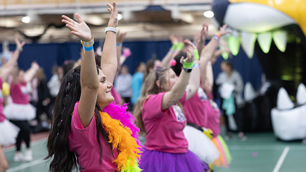 female students in pink shirts and colorful tutus wave their hands in the air as they dance