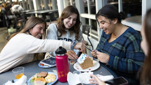Three students laugh as they build a gingerbread house together at a dining event