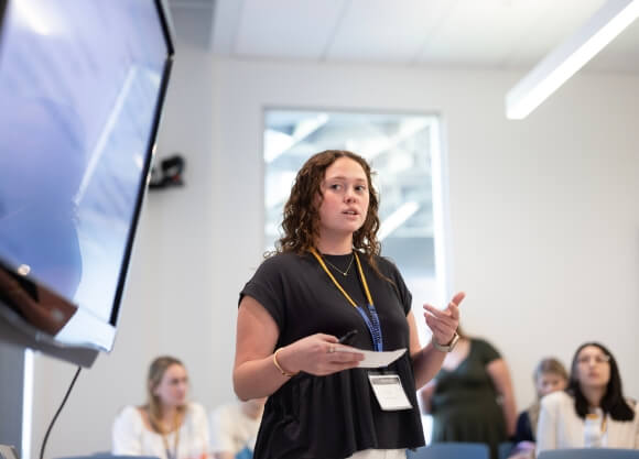 Photo of Madison Suk standing in front of a screen displaying her project while speaking to an audience of people in a classroom