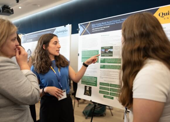 Photo of Lillian David and Sara Ottomanelli standing in front of poster and speaking to a female listener