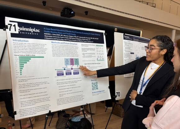 Photo of Noemia Chaloner standing in front of a poster and gesturing to a figure on the poster while a female is listening and looking at the poster