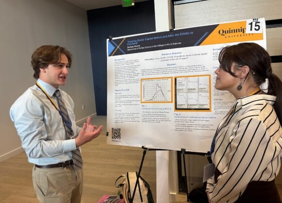 A photo of Zachary Pereira standing in front of poster and speaking to a female listener