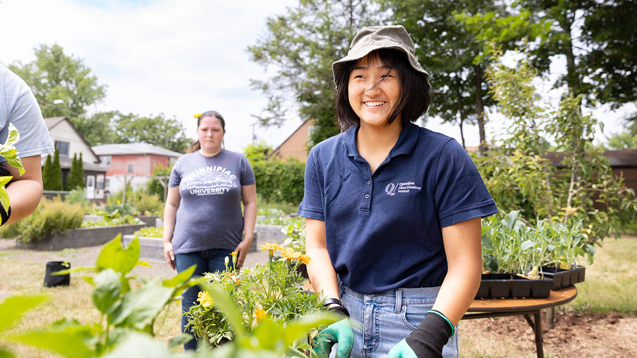 Quinnipiac student smiling with a bucket hat and gardening gloves on while planting