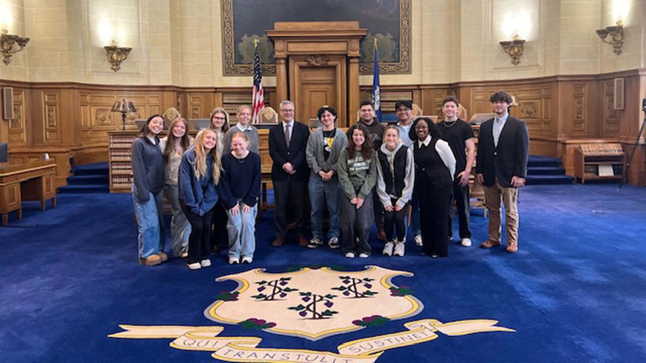 A group of 14 students and Justice Bright stands in the courtroom, on the carpet with the Connecticut seal.