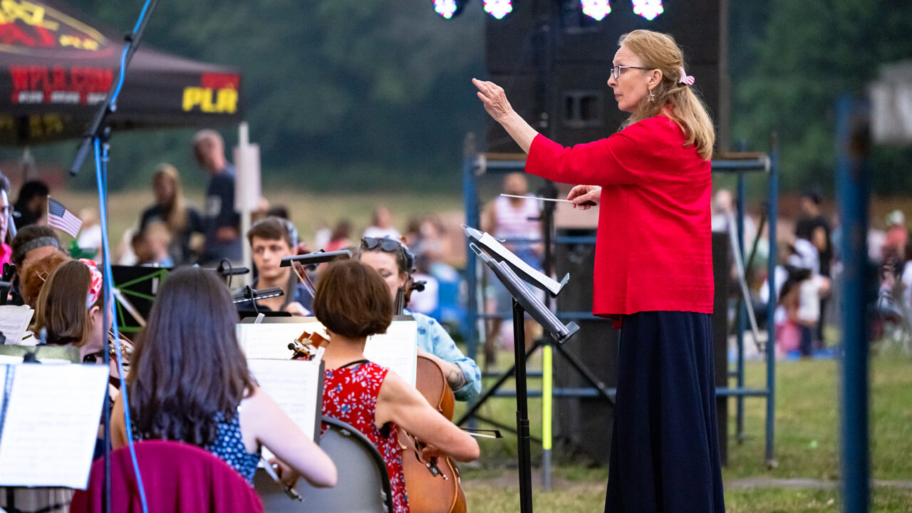 A conductor in a red shirt waves her baton in front of musicians on the Hamden town green