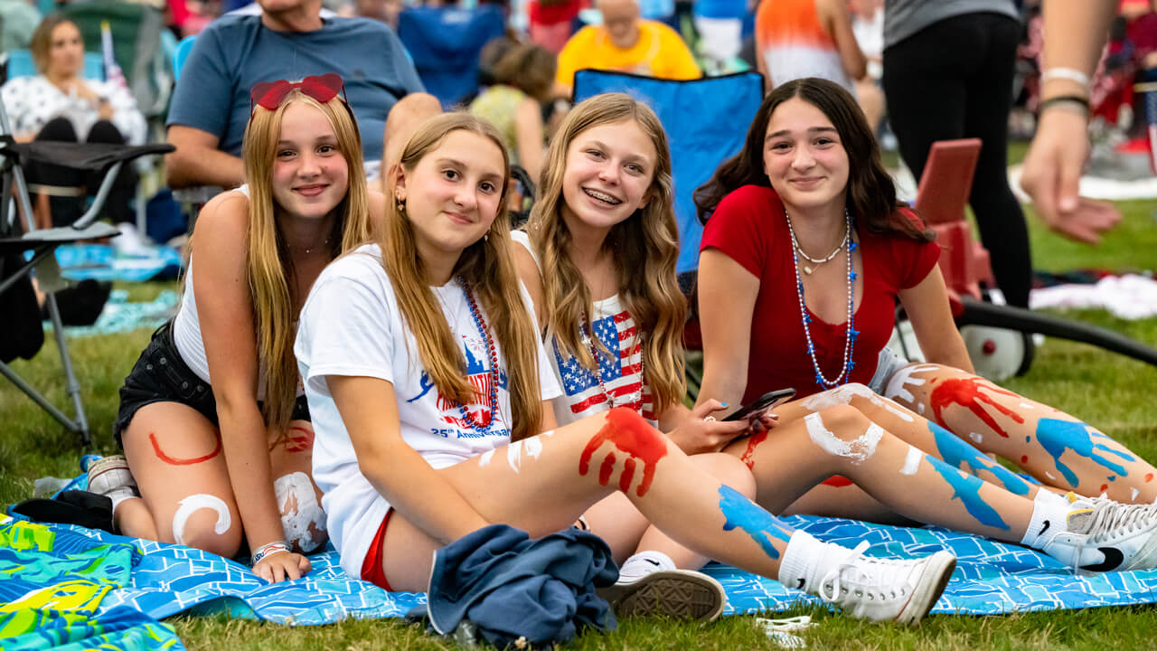 Four young people wear red, white and blue outfits and colorful accessories and smile together