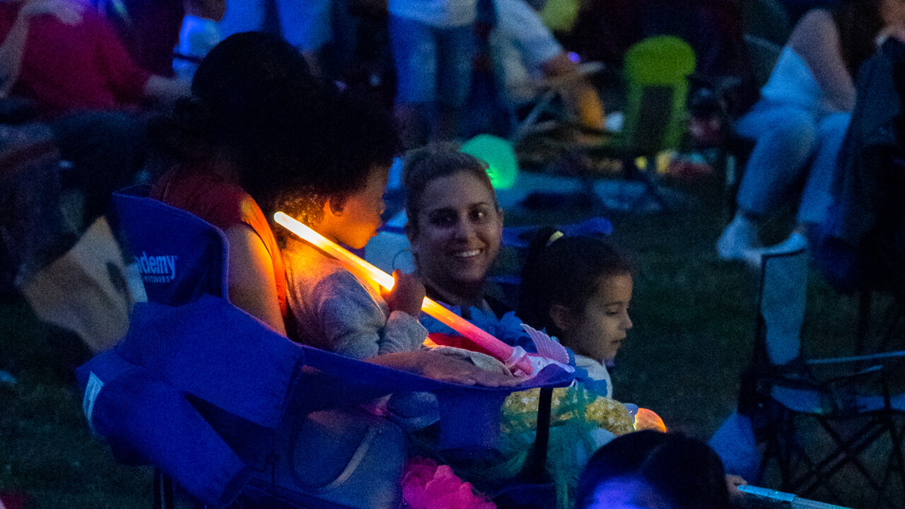 Families member smile at each other as their faces light up from glow sticks in the twilight