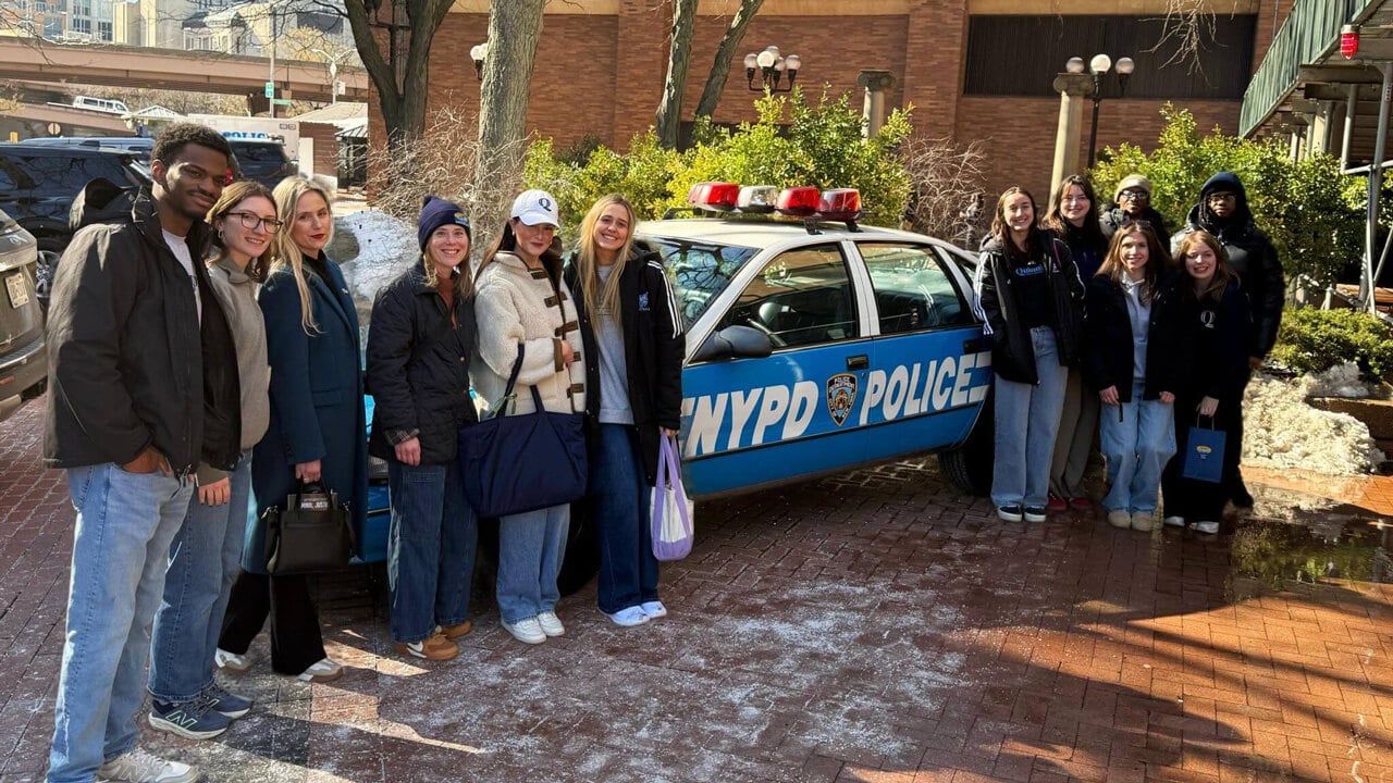 A group of 12 students and faculty members stand on either side of an NYPD police car.