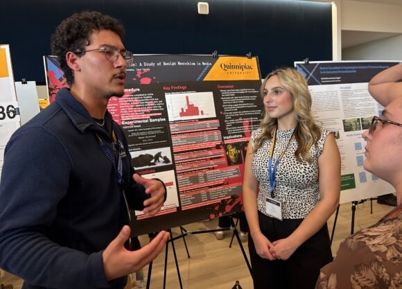 Photo of Mila Iuteri and Julien Mercado Bonanno standing in front of poster project and speaking to a female listener