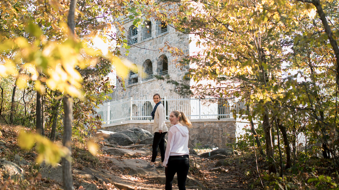 Two students hike up a rocky hill on an autumn day with the Sleeping Giant rock tower in the distance