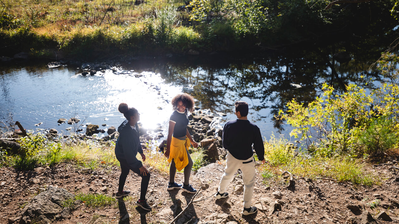 Three students walk along the bank of a river with the sun reflecting