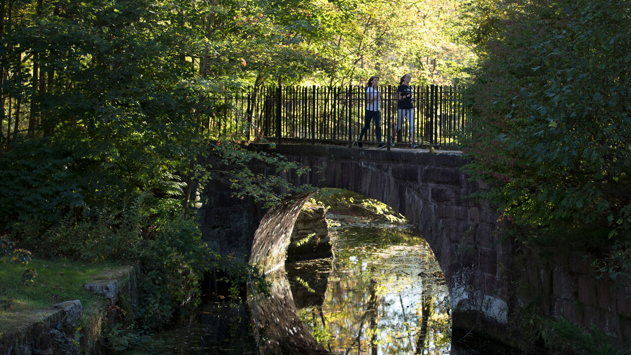 Two students jog across a stone arch bridge on a fall day