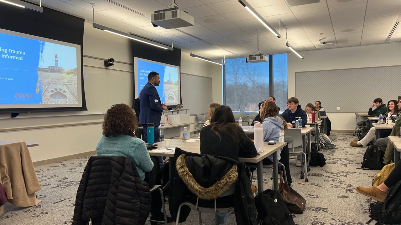 A speaker speaks in front of the class with a PowerPoint slide reading "Understanding Trauma and Trauma Informed Care."