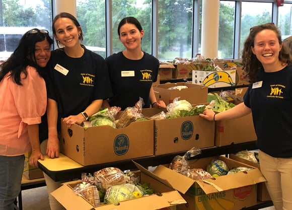 3 female students and 1 professor standing in front of boxes of food at a food bank