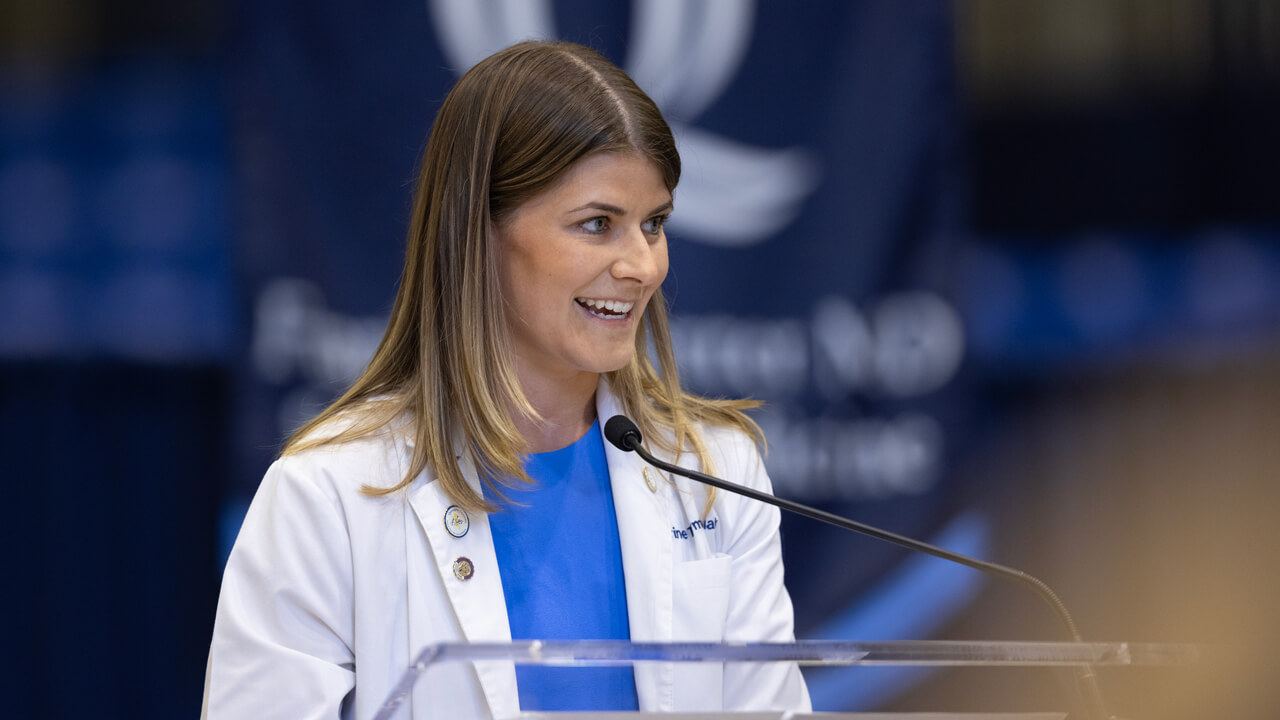 Medical student Katherine Tymbulak wears her white coat and addresses people from the podium