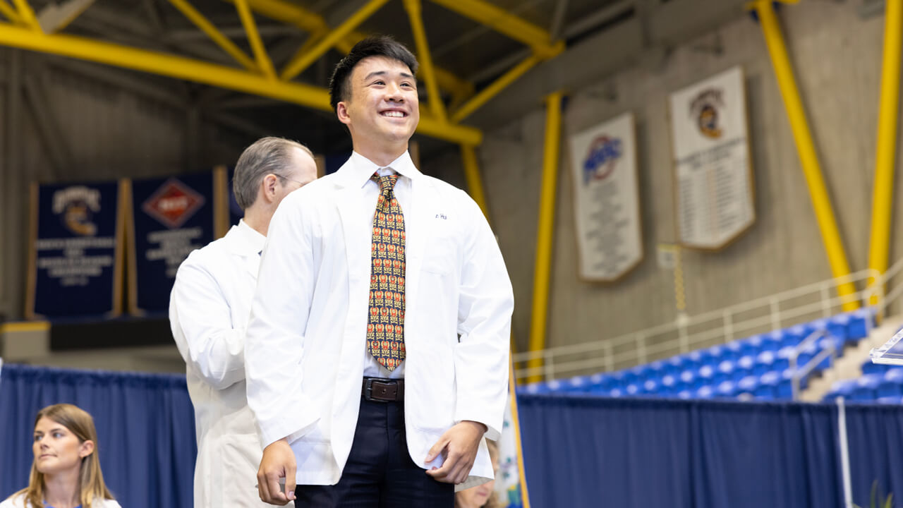 A student smiles broadly from stage as he wears his white coat