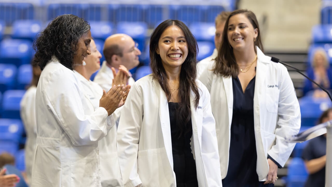 A student smiles broadly wearing her white coat while faculty members clap on stage