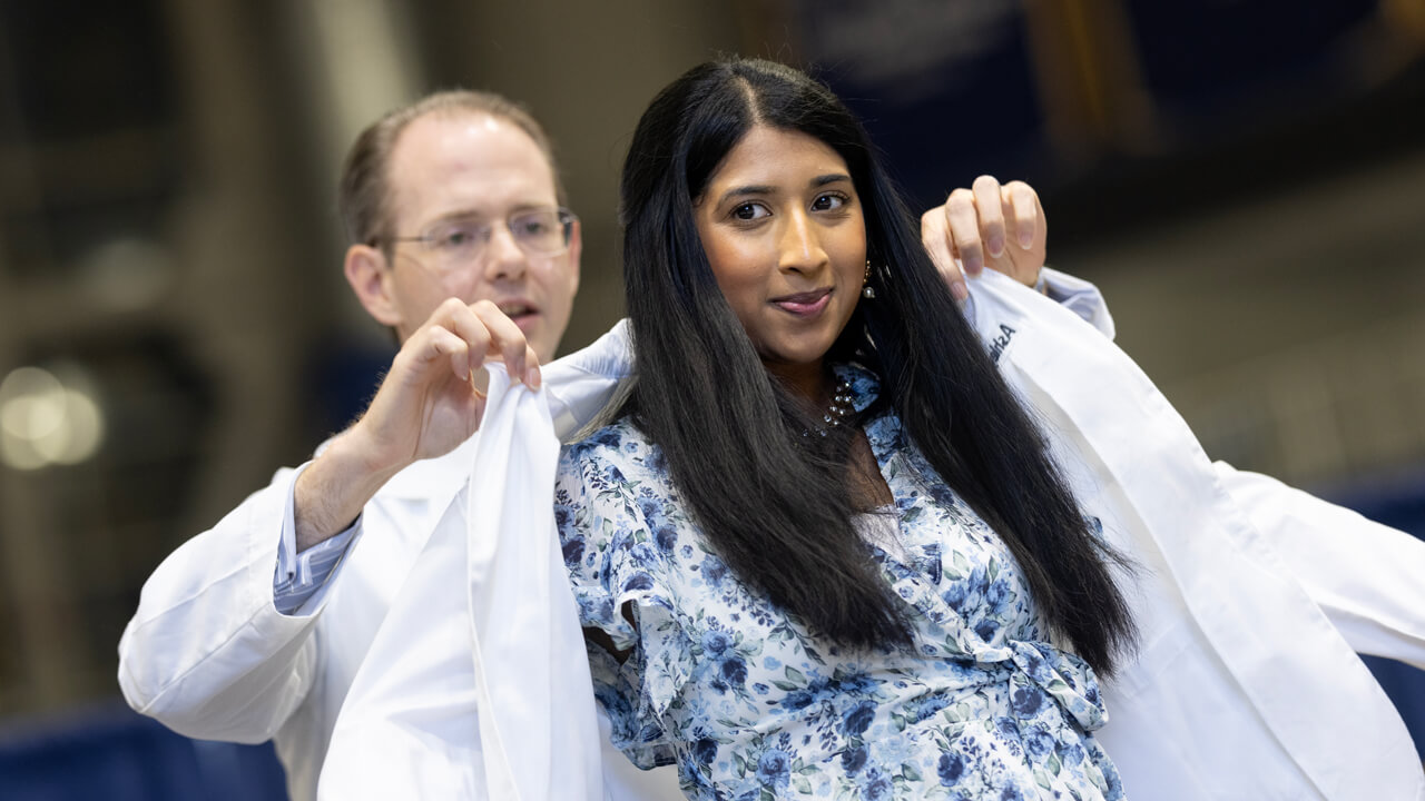A new medical student has her white coat lifted onto her shoulders by a faculty member