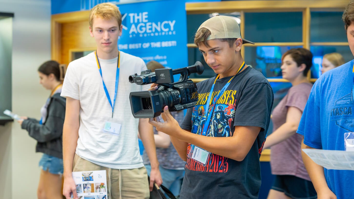 High school student wearing a baseball cap holds a UX-90 video camera, focusing intently while another student watches nearby during Quinnipiac University’s Movie Making Summer Camp.