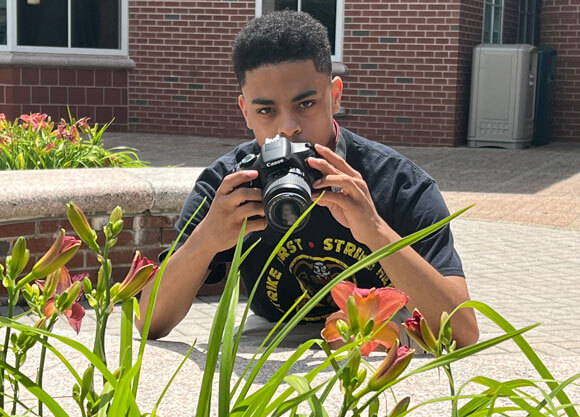 Prince Davenport taking pics with his camera lens behind plants and flowers