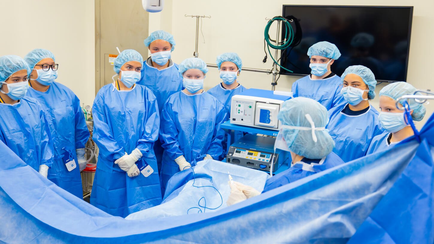 A group of students in full surgical scrubs and masks gather closely around a surgical table, observing a demonstration in a medical simulation lab.